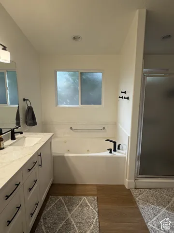 Bathroom featuring a white vanity with a light-toned stone countertop, recessed soaking tub, walk-in shower with a framed glass door, matte black fixtures, and wood-finish flooring