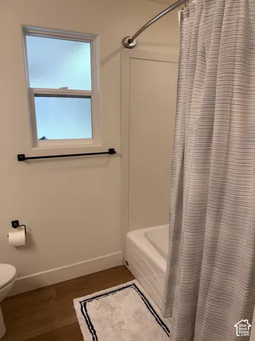 Bathroom featuring a white molded tub surround, frosted window, wood-finish flooring, and matte black fixtures including a towel bar and paper holder
