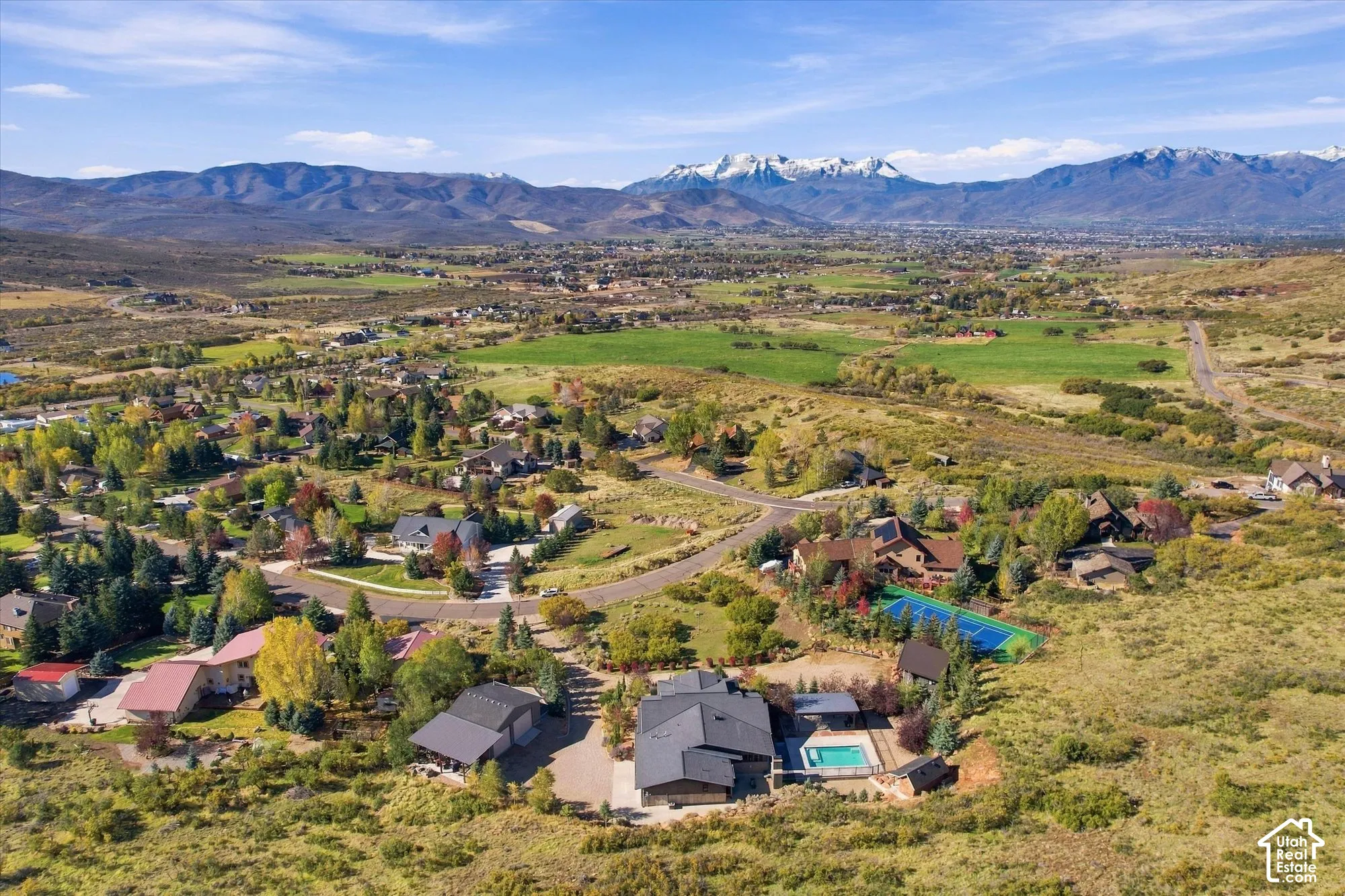 Aerial perspective of a property featuring an outdoor swimming pool, a private tennis court, and an attached multi-car garage, framed by expansive mountainous scenery