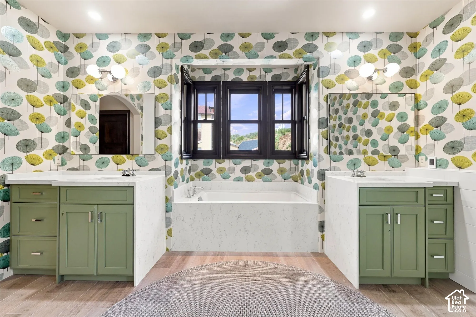 Dual vanities featuring sage green cabinetry, stone-finish countertops, and rectangular under-mount sinks, accompanied by a central soaking tub and wood-finish flooring