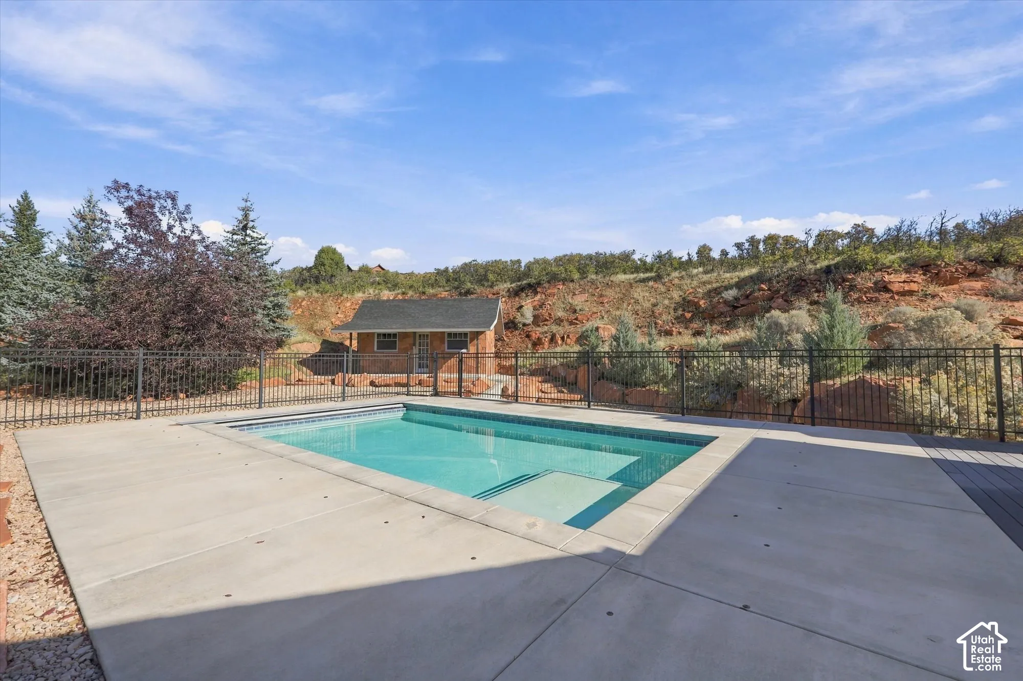 In-ground pool with integrated tanning ledge, expansive concrete decking, black metal perimeter fencing, and detached brick auxiliary structure