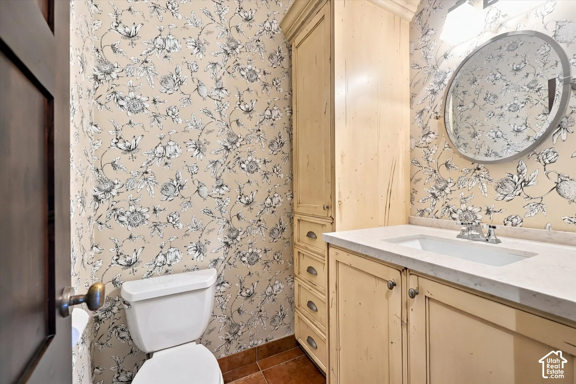Custom floor-to-ceiling built-in cabinetry, stone-finish countertop with an integrated rectangular sink, round wall-mounted mirror, and decorative floral wallpaper