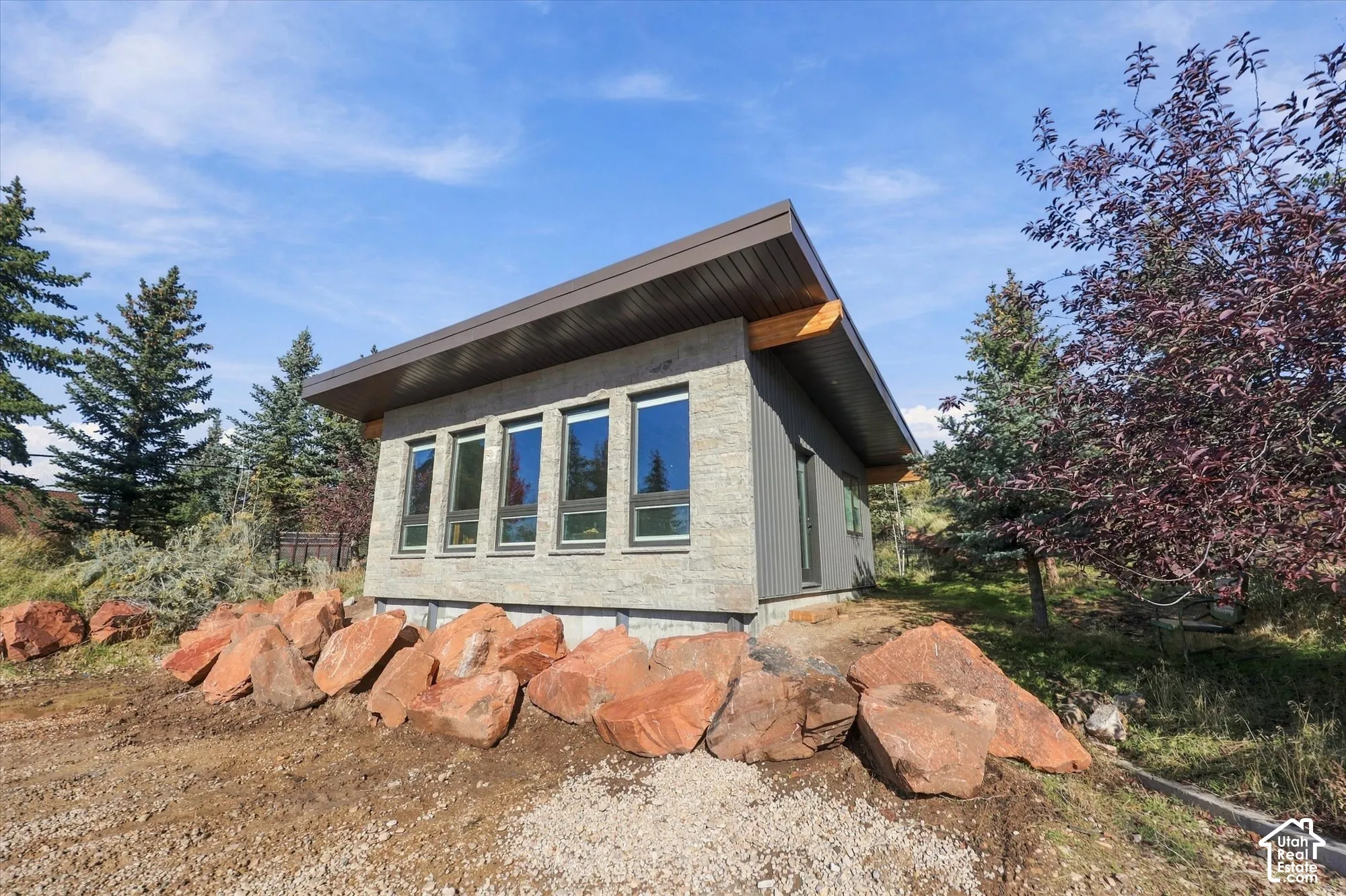 Modern exterior featuring a shed roof, vertical siding, stacked stone veneer, and oversized windows, set against a natural landscape with large boulder outcroppings