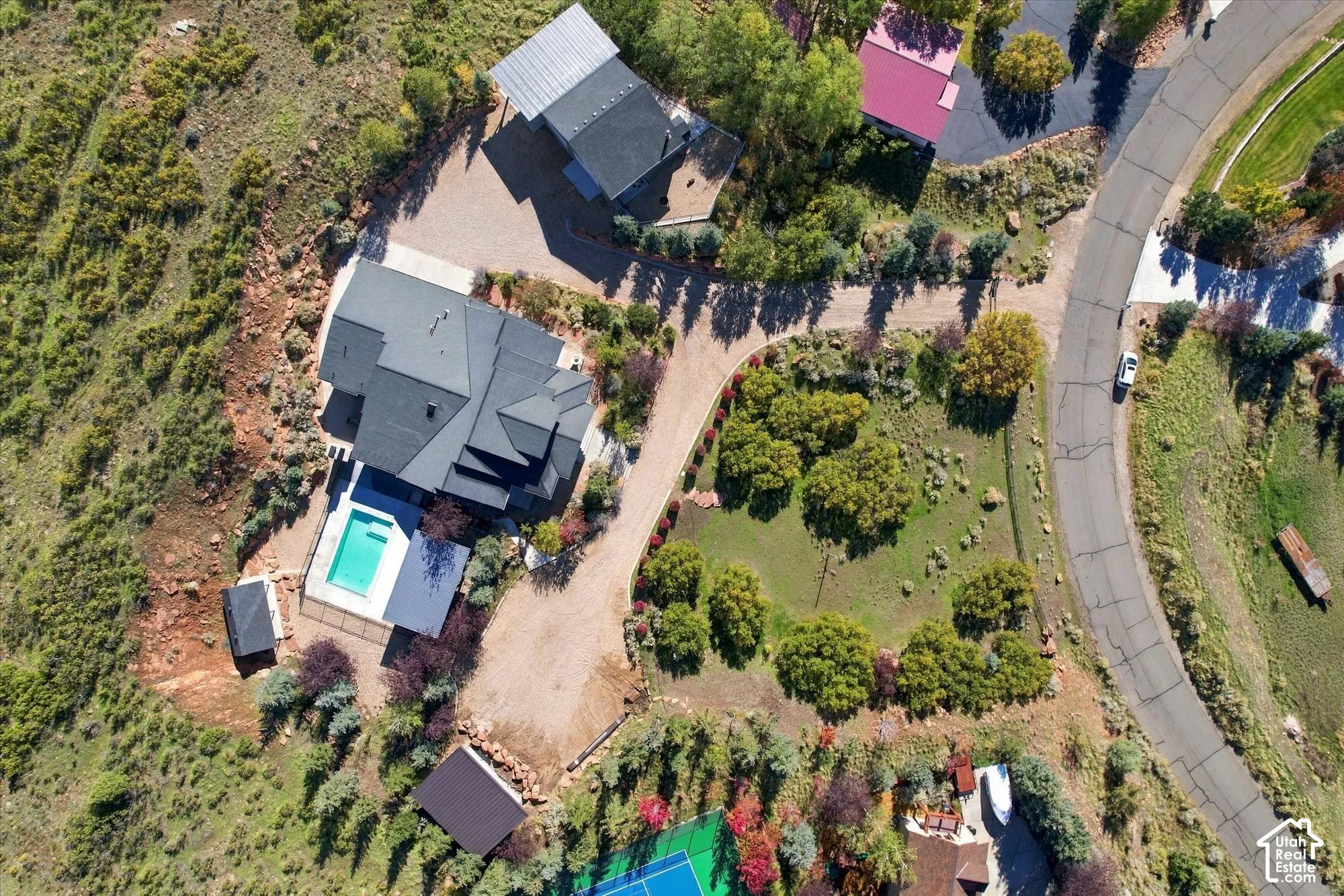 Aerial perspective of a hillside property featuring a multi-level residence with a dark-tone shingle roof, an in-ground swimming pool, a private tennis court, and an expansive graveled driveway