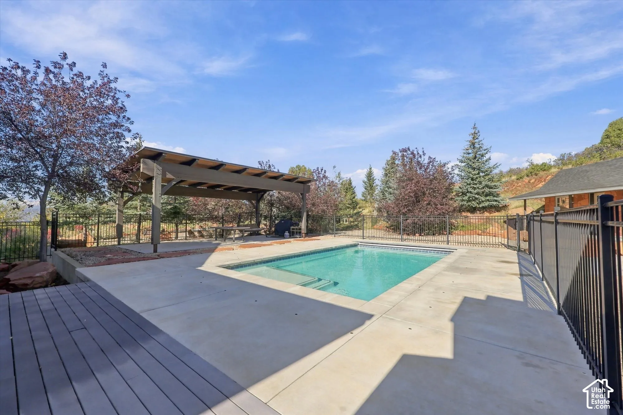 In-ground swimming pool with concrete decking, covered pergola, composite wood-finish decking, and black metal perimeter fencing
