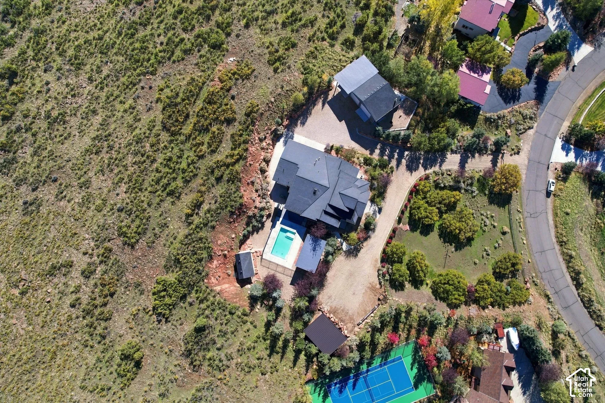 Aerial perspective of a property featuring an in-ground swimming pool, a private tennis court, expansive grounds, and a multi-level structure with a dark-toned roof