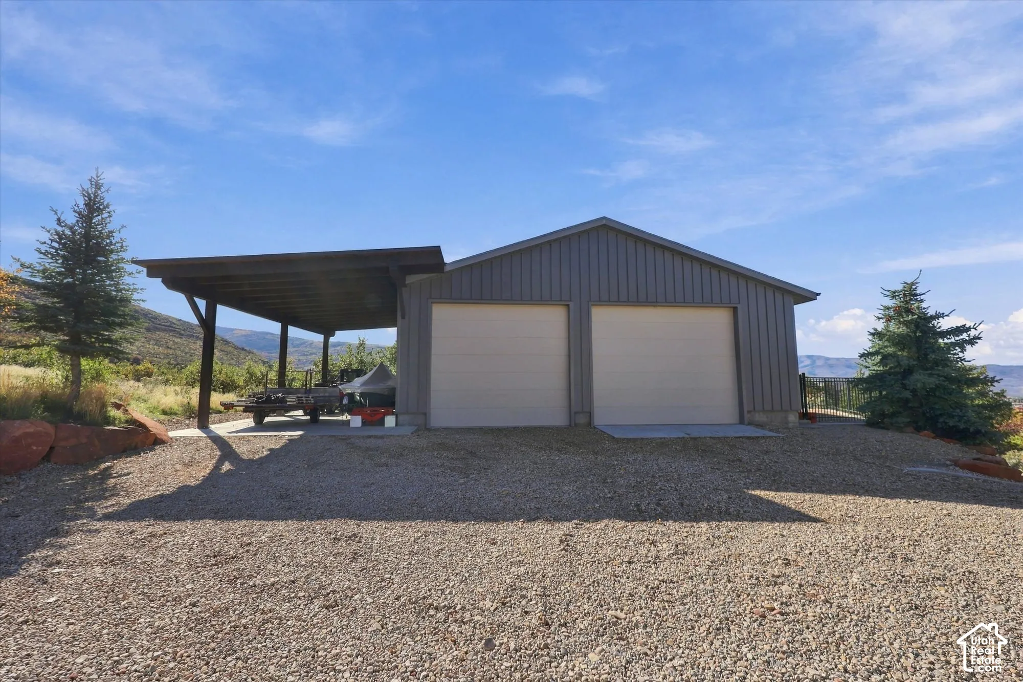 Detached metal-sided structure featuring two garage bays and a side-mounted covered carport, set on a gravel surface with mature evergreen landscaping