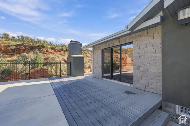 Rear patio featuring dual-material concrete and dark-toned wood-finish decking, exterior stone cladding, an outdoor fireplace, and a black metal perimeter fence