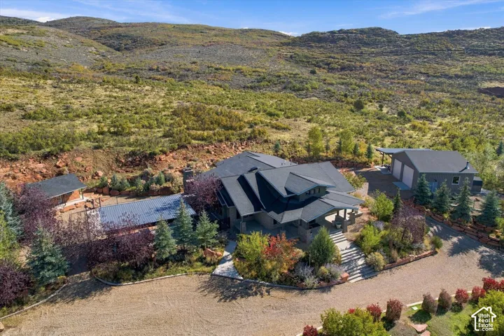 Multi-structure property featuring a primary residence with metal roofing, a detached garage, expansive gravel driveway, and mature landscaping set against a mountainous hillside backdrop