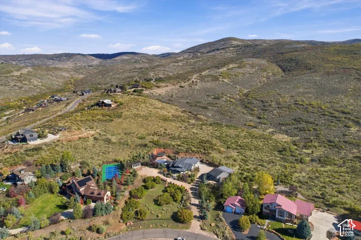 Aerial perspective of a hillside property featuring a tennis court, paved driveway, mature trees, and expansive mountainous terrain