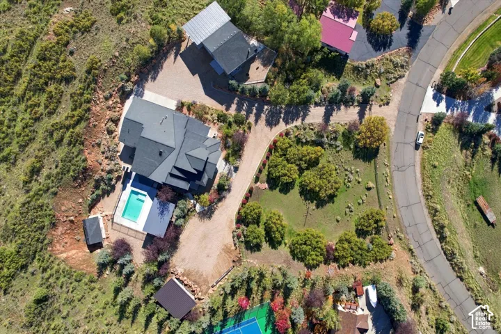 Aerial perspective of a hillside property featuring a multi-level residence with a dark-tone shingle roof, an in-ground swimming pool, a private tennis court, and an expansive graveled driveway