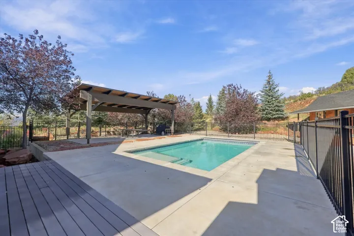 In-ground swimming pool with concrete decking, covered pergola, composite wood-finish decking, and black metal perimeter fencing