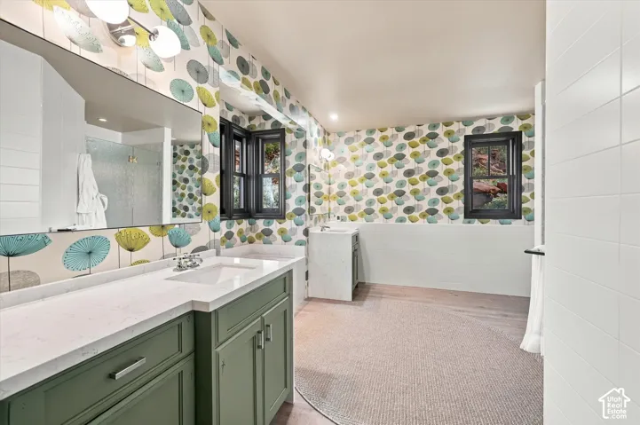 Bathroom featuring dual sage green cabinetry, stone-finish countertops, wall-to-wall mirrors, patterned wall coverings, and wood-finish flooring