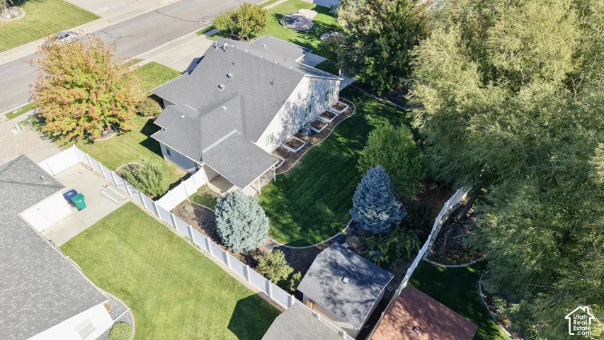 Aerial perspective featuring a manicured lawn, white vinyl perimeter fencing, mature trees, a detached storage shed, and stone-edged garden beds