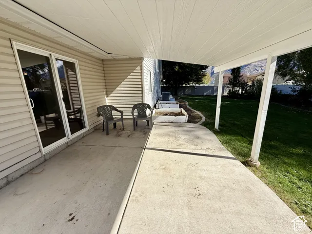 Covered concrete patio featuring horizontal vinyl siding, sliding glass doors, and a grass lawn with built-in raised garden beds