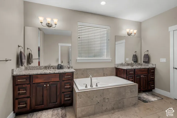 Dual dark wood vanities with stone-patterned countertops, large rectangular mirrors, recessed lighting, and a drop-in soaking tub encased in neutral stone-finish tile