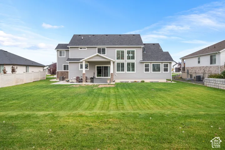 Rear exterior featuring grey horizontal siding, a covered concrete patio, stone-accented columns, expansive grass lawn, and white vinyl privacy fencing
