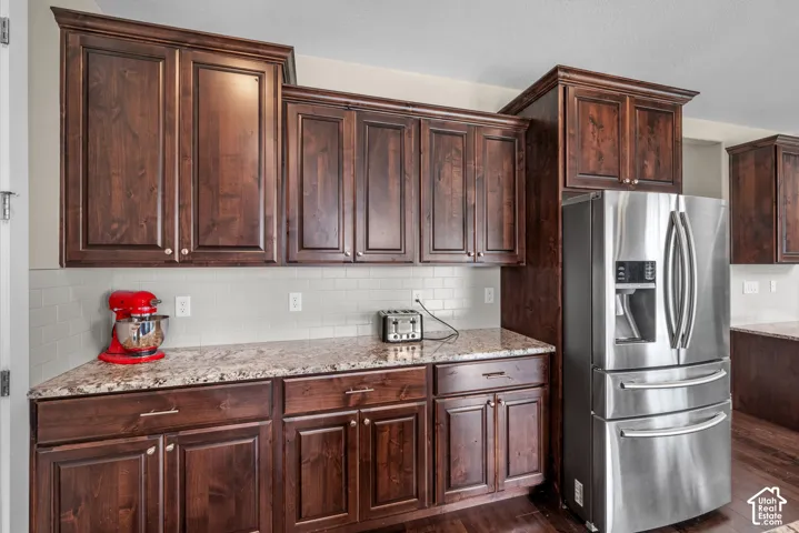 Dark wood cabinetry with crown molding, stone-finish countertops, subway tile backsplash, and stainless steel refrigerator