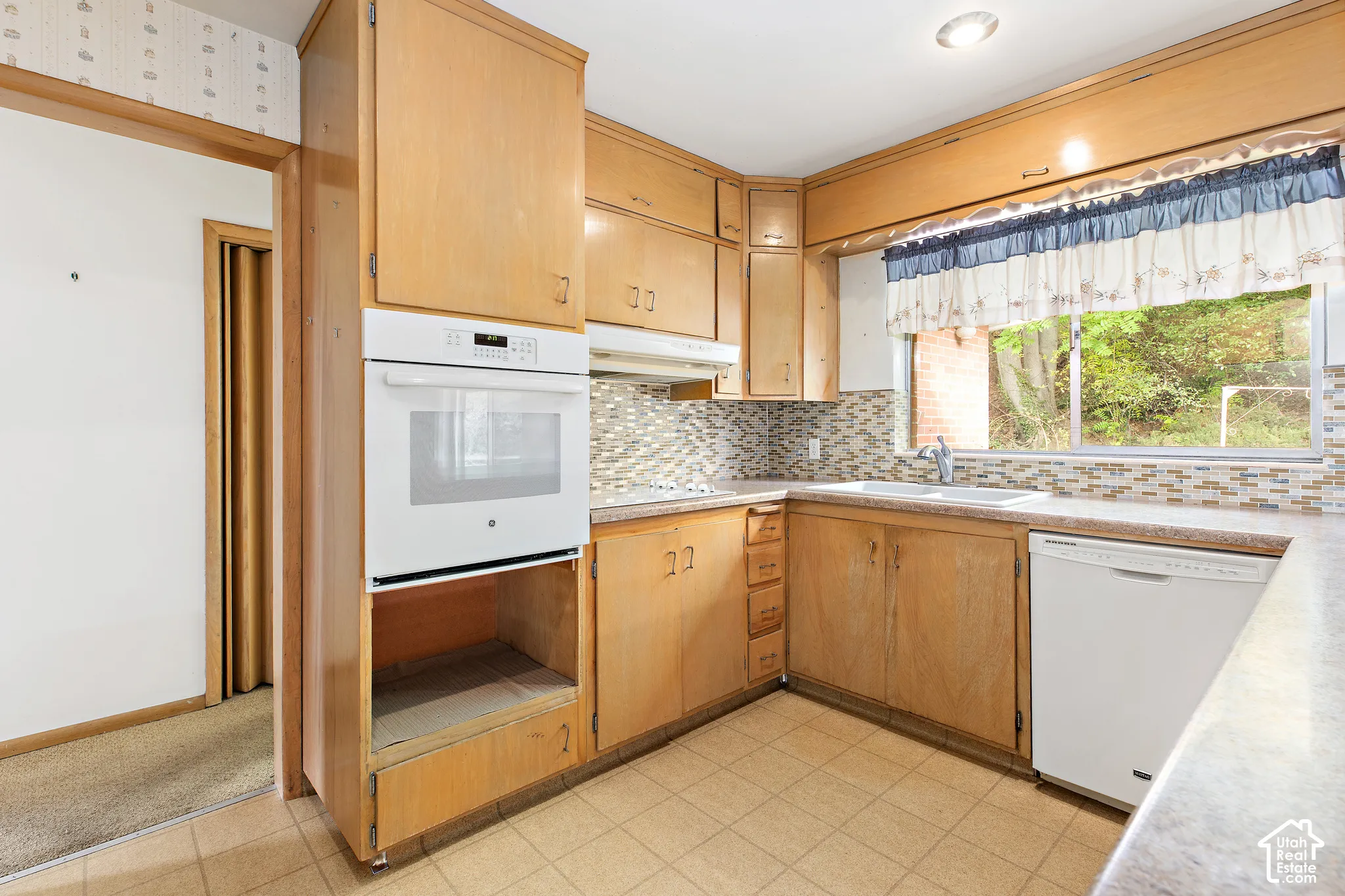 Kitchen featuring wood cabinetry, built-in wall oven, multi-toned mosaic tile backsplash, double-basin sink with chrome faucet, and tiled flooring