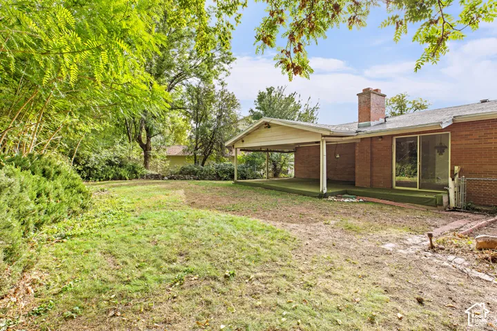 Brick exterior with a covered patio, masonry chimney, and expansive lawn area featuring mature trees