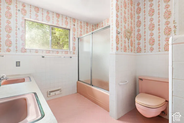 Bathroom featuring a dual-sink vanity with a solid-surface countertop, a porcelain tub with a sliding glass door, ceramic wall tiling, and pink-toned fixtures