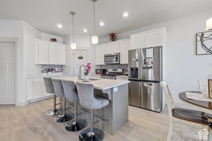 Kitchen featuring white shaker-style cabinetry, quartz countertops, a multi-tone tile backsplash, stainless steel appliances, and a center island with a built-in sink