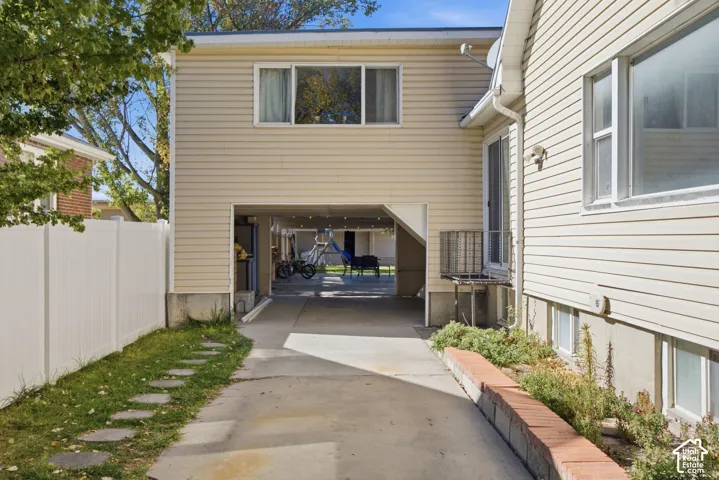 Exterior featuring beige horizontal siding, a concrete driveway leading to an open-air carport, a white vinyl privacy fence, and a tiered brick garden border