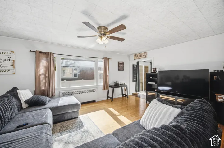 Living space featuring wood-finish flooring, a large picture window, a central ceiling fan with integrated lighting, and decorative radiator cover