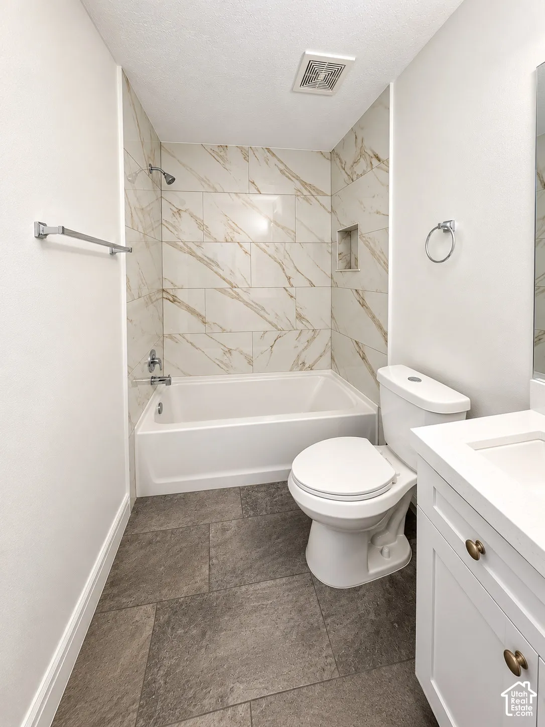 Full bathroom featuring a tub-shower combination with floor-to-ceiling veined tile surround, recessed shower niche, white vanity cabinetry with an integrated countertop, and large-format tile flooring