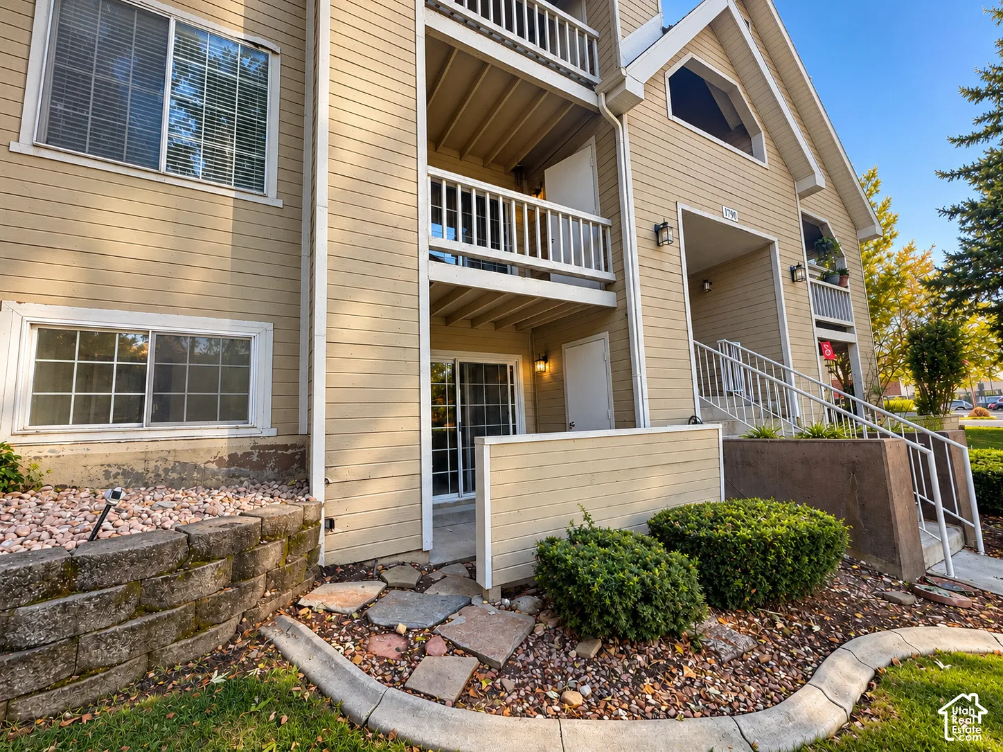 Multi-story exterior featuring horizontal siding, private balconies with white railings, sliding glass door entry, and a stone-block retaining wall