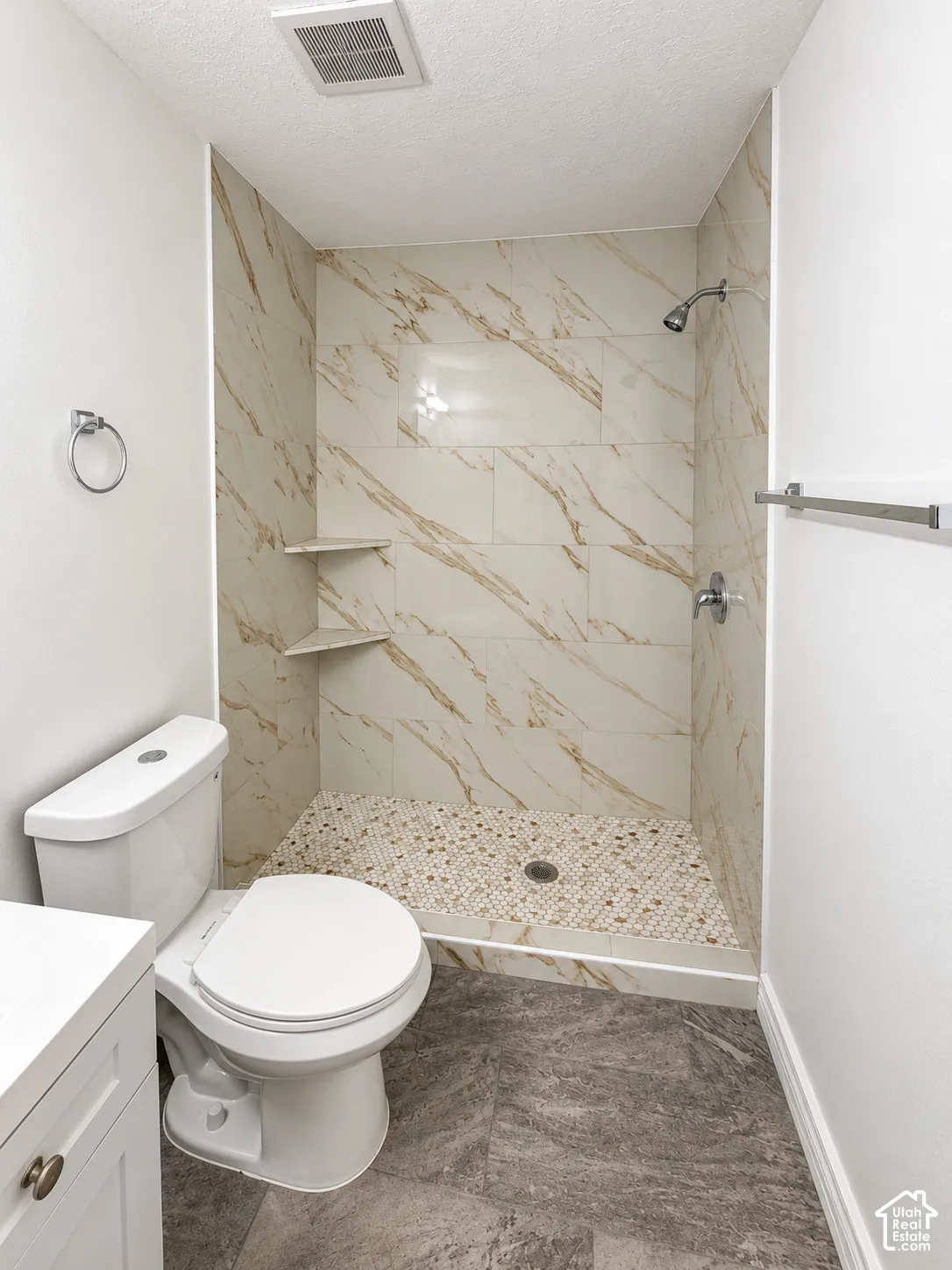 Bathroom featuring stone-finish tile flooring, walk-in shower with large-format patterned wall tile, pebble-texture shower floor, built-in corner shelving, and polished chrome fixtures