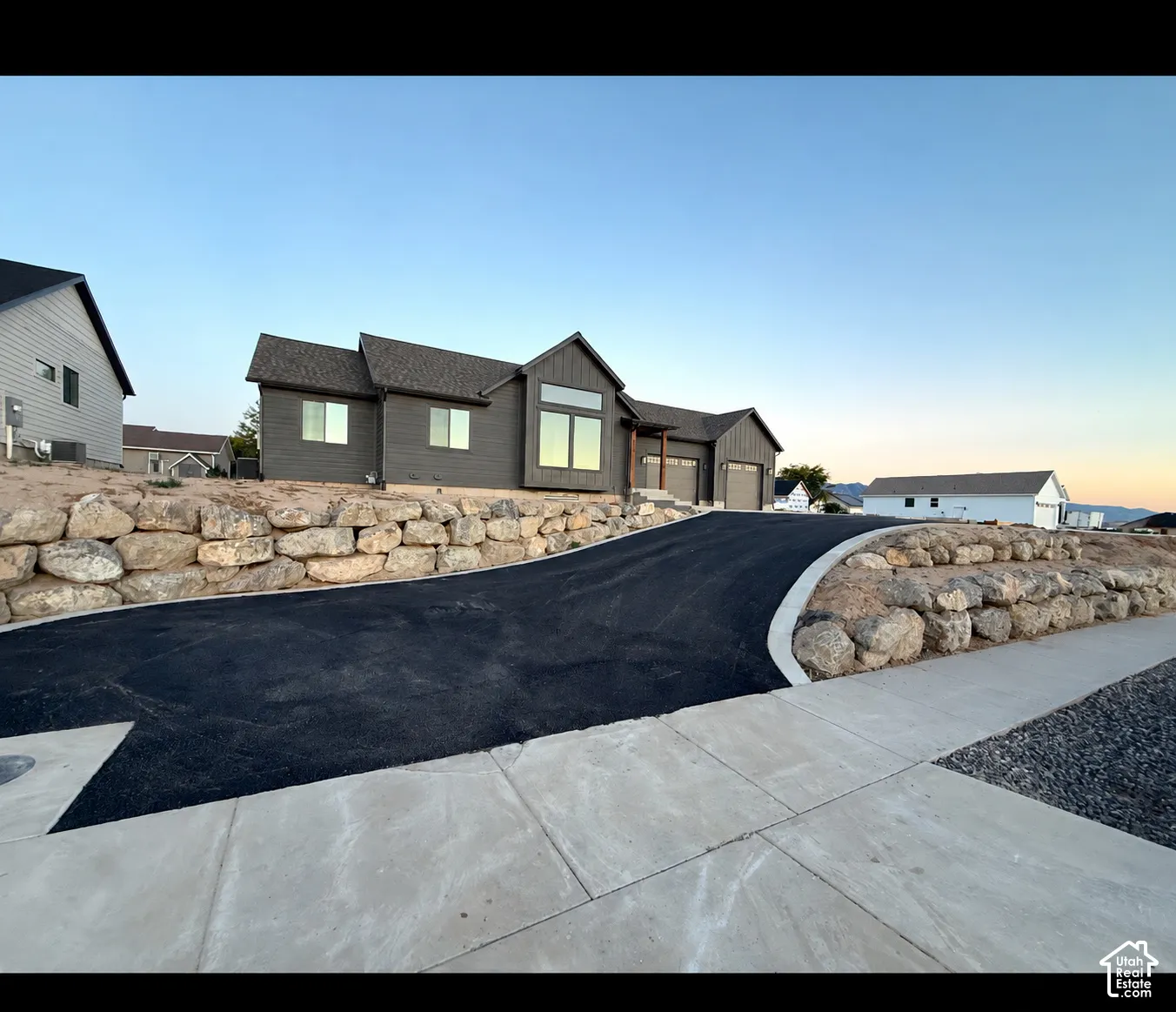 Modern residence with dark-toned horizontal siding, asphalt driveway, stone retaining walls, and multi-car garage