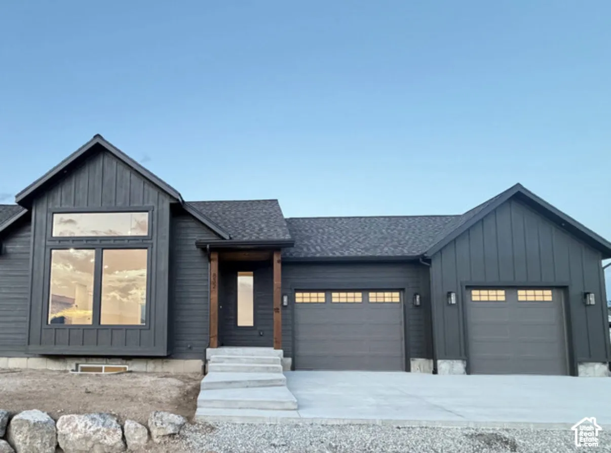Dark vertical siding exterior with wood-post porch detailing, large picture windows, concrete driveway, and attached two-car garage