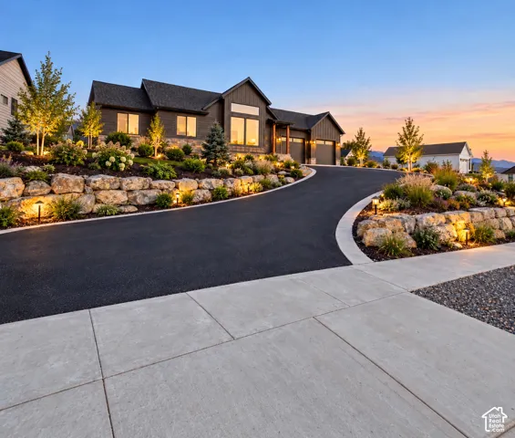Exterior facade featuring board and batten siding with stone accents, tiered rock retaining walls, paved driveway, and mountain backdrop