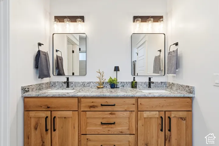 Double vanity featuring natural wood-finish cabinetry, stone countertop with integrated dual sinks, matte black hardware, dual rectangular mirrors, and multi-light vanity fixtures