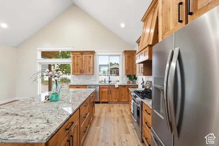 Kitchen featuring vaulted ceilings, wood-finish cabinetry, an oversized stone-surface island, stainless steel appliances, and wood-finish flooring