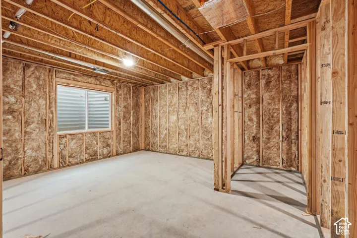 Unfinished interior space featuring exposed wood framing, installed fiberglass batt insulation, concrete flooring, and an egress window with a corrugated metal window well