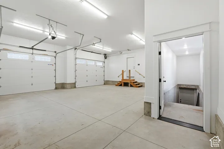 Spacious three-car garage featuring finished white walls, concrete flooring, dual overhead lighting, and an integrated staircase with wooden handrails