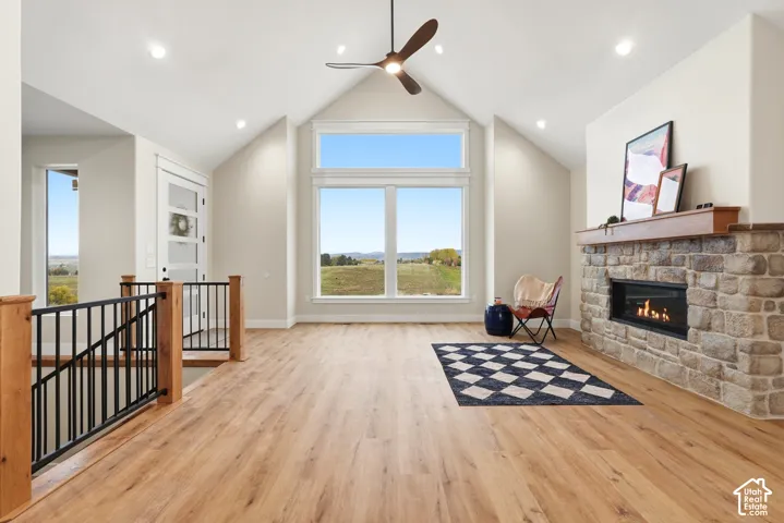 Vaulted ceiling featuring a central modern fan, light-toned wood-finish flooring, floor-to-ceiling stone-clad fireplace with a natural wood mantel, and large-scale windows with metal railings
