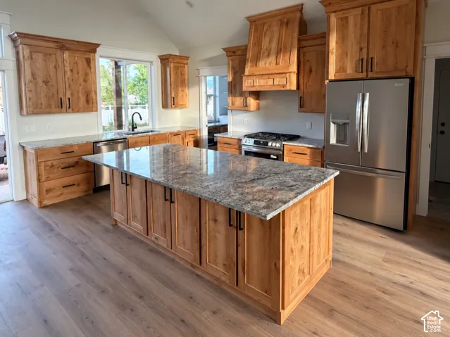 Kitchen featuring custom wood cabinetry, an expansive center island with a stone-finish countertop, stainless steel appliances, a vaulted ceiling, and wood-finish flooring