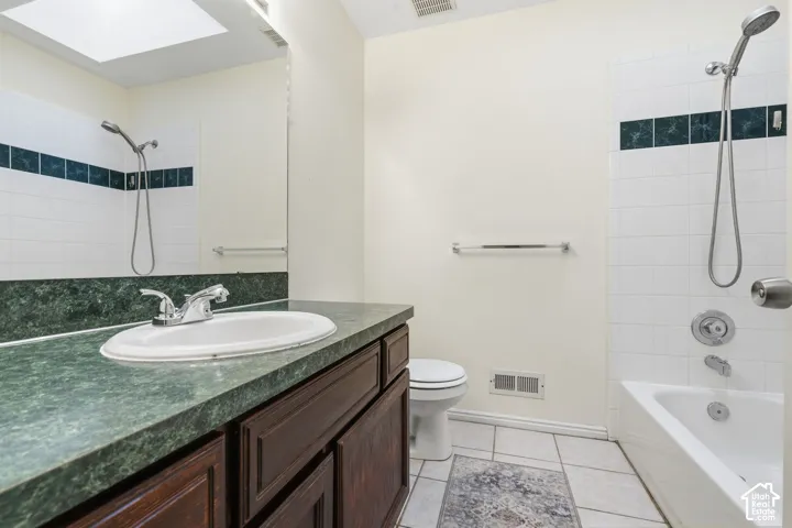 Bathroom featuring a wood-finish vanity with a stone-texture countertop, integrated sink, tile flooring, and a shower-tub combination with decorative accent tiling