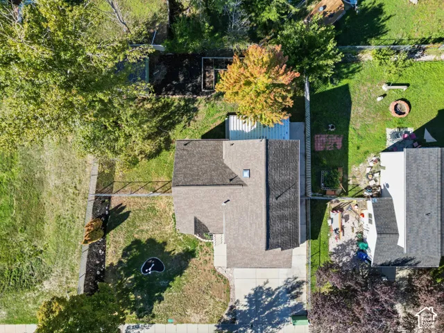 Aerial perspective featuring a multi-pitched shingle roof, concrete driveway, fenced yard, and established trees