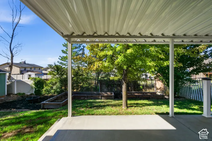 Covered concrete patio featuring a metal roof structure, built-in timber garden beds, grass lawn, and mature trees
