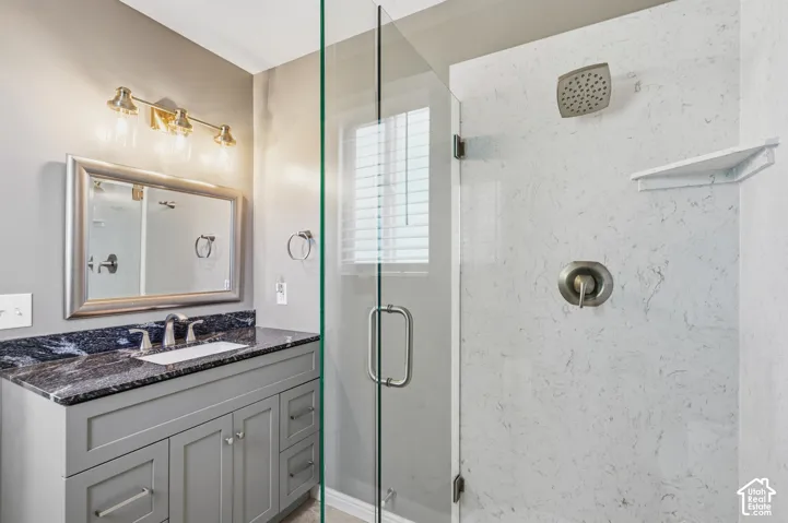 Bathroom featuring a grey vanity with dark stone countertop, undermount sink, glass-enclosed shower with stone-patterned surround, and three-light wall fixture