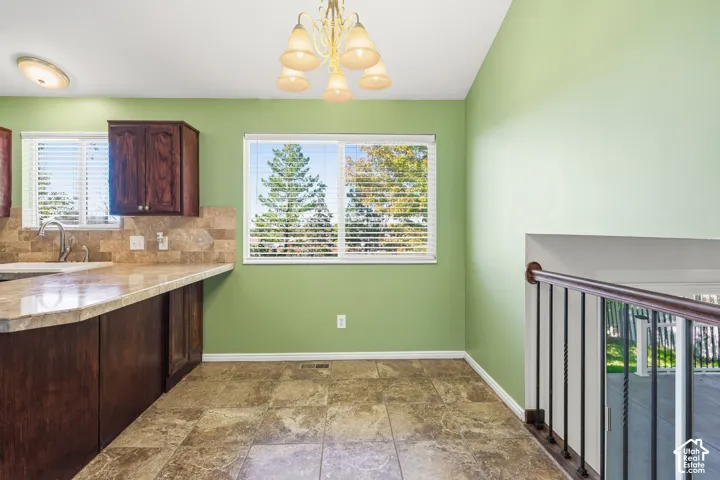 Kitchen area featuring dark wood cabinetry, natural stone-finish backsplash, neutral-tone countertop, tile flooring, and metal railing with a wood handrail