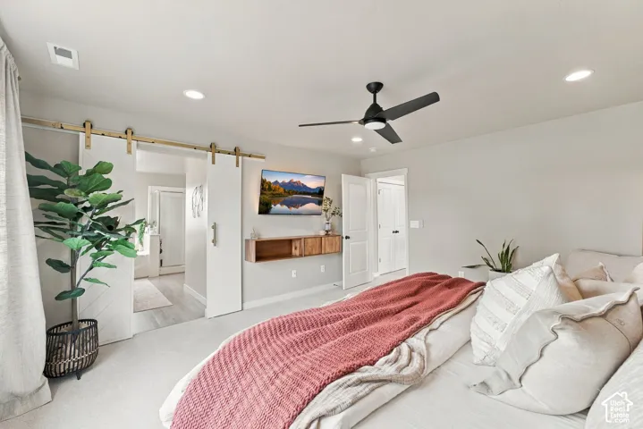 Room featuring white barn-style sliding doors with gold-finish hardware, recessed ceiling lighting, a matte black ceiling fan, and a wall-mounted wooden floating shelf