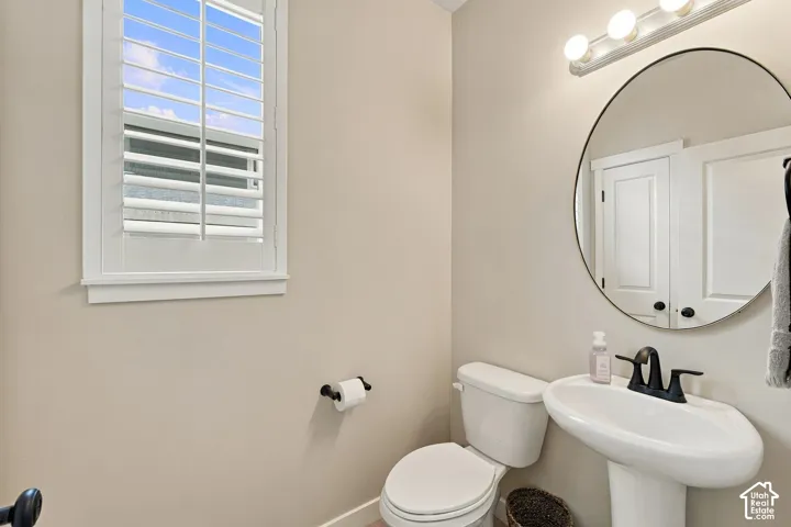 Powder room featuring a pedestal sink, matte black faucet, round wall-mounted mirror, three-light vanity fixture, and window with plantation shutters