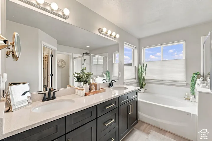 Dual-sink vanity with matte black fixtures, stone-surface countertop, full-width vanity mirror, deep soaking tub, and wood-finish flooring