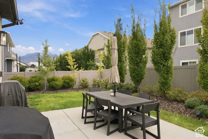 Rear yard featuring a concrete patio, manicured lawn, perimeter fencing, mature landscaping, and mountain backdrop