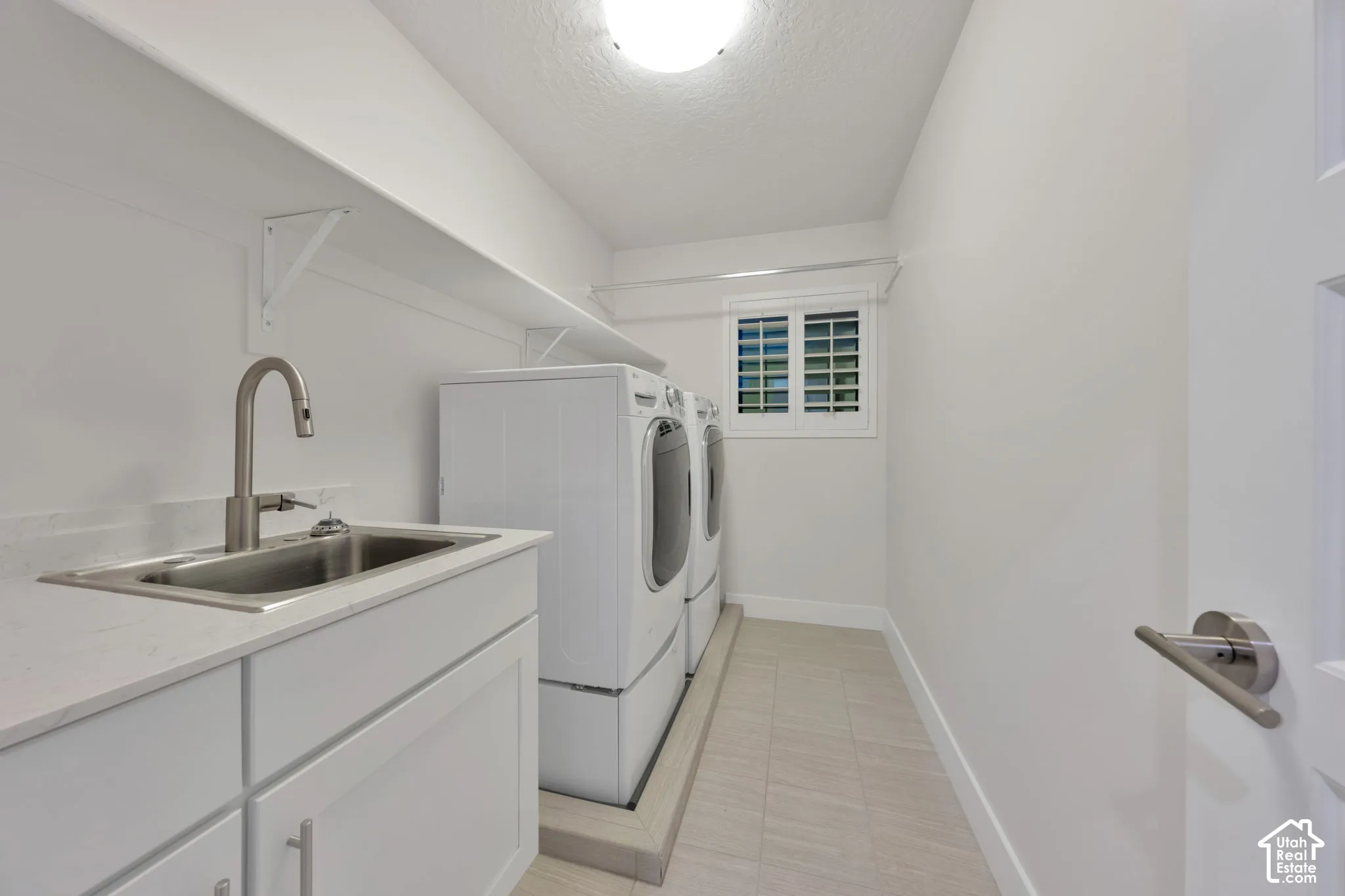Laundry room featuring white cabinetry, a built-in stainless steel sink with a high-arc faucet, tile flooring, and plantation shutters